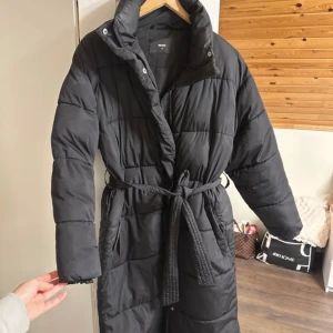 Svart lång pufferjacka från Bik Bok - Säljer en svart lång pufferjacka från Bik Bok med bälte i midjan. Jackan har hög krage, långa ärmar och quiltad design. Perfekt för kyliga dagar och enkel att styla med olika outfits.