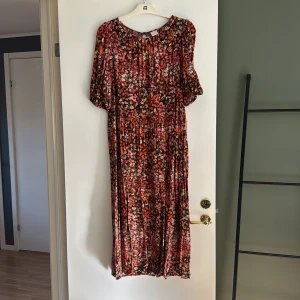 Blommig långklänning med kort ärm - Säljer en långklänning med färgstarkt blommönster i rött, rosa, vitt och grönt på svart botten. Klänningen har korta ärmar, rund halsringning och knäppning bak i nacken. Perfekt för dig som gillar romantiska och färgglada plagg.