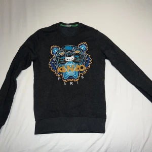 Svart sweatshirt från Kenzo med tigerbroderi - Säljer en svart sweatshirt från Kenzo med stort färgglatt tigerbroderi och texten 'KENZO PARIS' på bröstet. Tröjan har rund hals och långa ärmar. Perfekt statement-plagg för dig som gillar streetwear.
