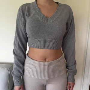 Grå croppad stickad tröja med v-ringning - Säljer en grå croppad stickad tröja med v-ringad halsringning och långa ärmar. Tröjan har bred ribbad kant nedtill och vid ärmsluten, vilket ger en snygg och modern look.
