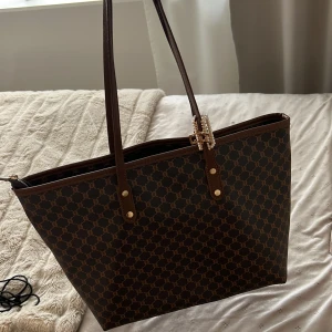 Brun axelväska med mönster - Snygg brun axelväska med klassiskt mönster och långa handtag. Väskan har guldfärgade detaljer och dragkedja upptill. Perfekt för att bära med sig allt man behöver under dagen.