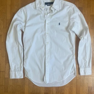 Vit skjorta från Ralph Lauren - Klassisk vit skjorta från Ralph Lauren med broderad logga på bröstet. Slim fit-modell med långa ärmar och knappar framtill. Perfekt för en stilren och clean look. Tillverkad i bomull för en skön känsla.