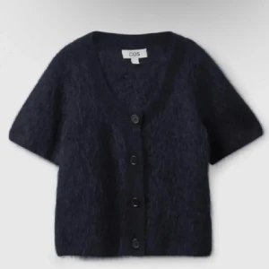 Svart kofta med kort ärm från COS - Säljer en svart, fluffig kofta från COS med korta ärmar och v-ringning. Koftan har fyra knappar framtill och är perfekt för lager-på-lager. Enkel och stilren design som passar till mycket.