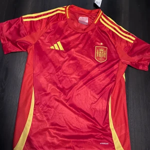 Spaniens landslagströja från Adidas - Röd kortärmad fotbollströja från Adidas med Spaniens landslagsmärke på bröstet och gula detaljer längs axlar och sidor. Klassisk rund hals och tillverkad i lätt, ventilerande material. Perfekt för fotbollsträning eller att bära som supporter.