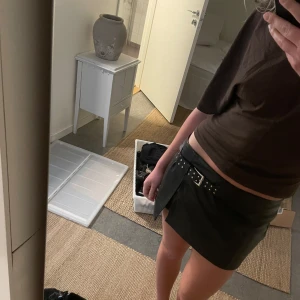 Svart skinnkjol med bälte - Säljer en svart kort skinnkjol med bälte och silverdetaljer. Från Bershka med inbyggda kjol i storlek 38