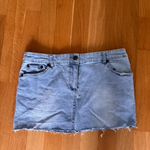 Ljusblå jeanskjol från OJARE - Ljusblå jeanskjol med rå kant nertill och klassiska fem fickor. Kjolen har knapp och dragkedja framtill samt dekorativa sömmar på bakfickorna. Perfekt för en avslappnad stil.