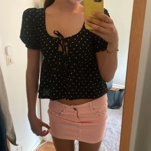 Svart blus med vita prickar - Säljer en svart blus med vita prickar och kort puffärm. Blusen har knytning framtill och en lätt v-ringning. Perfekt för varma dagar och ger en söt och trendig look.