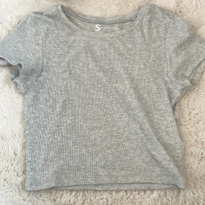 Grå ribbad croppad t-shirt från newyorker - Säljer en ljusgrå croppad t-shirt från newyorker. Den kommer ej till användning pga den är för kort enligt min smak