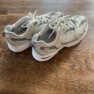 Vita och grå New Balance sneakers - Säljer ett par fräscha New Balance sneakers i vitt och grått med klassisk N-logga på sidan. Skorna har snörning och en bekväm, platt sula som passar perfekt till vardags. Stilren design med mesh och syntetmaterial för bra andningsförmåga.