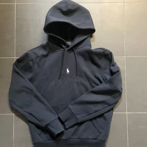 Mörkblå hoodie från Polo Ralph Lauren - Mörkblå hoodie från Polo Ralph Lauren med klassisk huva och dragsko. Tröjan har en broderad vit logga på bröstet och en stor magficka. Perfekt för en avslappnad stil.