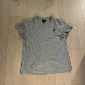 Grå t-shirt från Lyle & Scott Junior - Säljer en stilren grå t-shirt från Lyle & Scott Junior med klassisk rund hals och korta ärmar. T-shirten har en broderad gul logga på bröstet och är tillverkad i mjuk bomull. Perfekt för en avslappnad look.