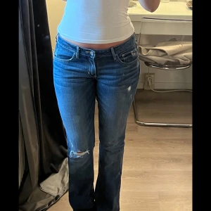 Blå bootcut jeans från Hollister - Säljer ett par blå bootcut jeans från Hollister med låg midja. Jeansen har slitningar och hål vid knät samt klassiska broderade bakfickor. Perfekta för dig som gillar en avslappnad och trendig look.