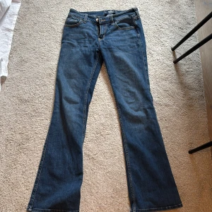 Blå bootcut jeans från Hollister - Säljer ett par klassiska blå bootcut jeans från Hollister med låg midja och mjukt stretchigt material. Jeansen har fem fickor, snygga sömmar och läderpatch med logga bak. Perfekta för dig som gillar en tidlös jeansstil.