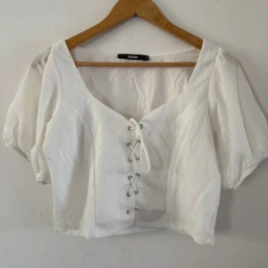 Vit snörad blus från Bik Bok - Säljer en vit blus från Bik Bok med puffiga korta ärmar och snygg snörning framtill. Toppen har en croppad passform och är tillverkad i ett lätt bomullsmaterial. Perfekt för en trendig och luftig look.