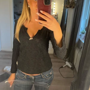 Svart långärmad topp med rosett - Säljer en svart långärmad topp med diskret mönster och liten söt rosett framtill vid halsringningen. Toppen har en lätt croppad passform och v-ringad hals. Perfekt att styla med jeans eller kjol.