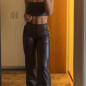 Bruna skinnbyxor med bootcut - Säljer ett par bruna skinnbyxor med bootcut samt tillhörande topp. Byxorna har en slät framsida med dragkedja och sitter tight upptill men släpper vid benen. Korsett toppen har en dragkedja baktill.