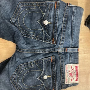 True Religion jeans med kontrastsömmar - Säljer ett par klassiska blå jeans från True Religion med tydliga kontrastsömmar och stora bakfickor med signatursöm. Jeansen har låg midja och raka ben, samt detaljer i silverfärg och orange. Perfekta för dig som gillar streetwear och vill sticka ut.