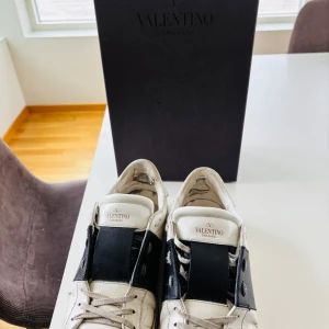 Valentino Garavani - Säljer ett par vita sneakers från Valentino Garavani med bred svart läderdetalj över mitten. Skorna har rund tå, platt sula och klassisk snörning. Kommer med originalkartong och tillbehör. Perfekta för dig som gillar stilrena och exklusiva sneakers. Skorna är i skick 7/10 men är inte hål så det syns eller märks vid användning. Priset kan diskuteras vid snabb affär.