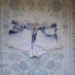 Vita jeansshorts med spetsdetaljer - Säljer ett par vita jeansshorts med ljusblå tvätt och blommiga spetsdetaljer längs bensluten. Shortsen har klassisk femficksdesign, bälteshällor och knappgylf. Perfekta för sommaren!