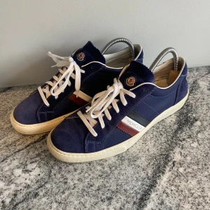Mörkblåa moncler skor strl 42 men passar 43 - Snygga blå sneakers från Moncler med vita snören och röd-vit-blå rand på sidan. Skorna har rund tå, platt sula och Moncler-logga på plösen. Materialmix av mocka och skinn ger en exklusiv känsla. Med mocka där fram och bak. Pris kan diskuteras. 