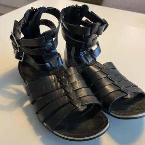 Svarta sandaler med spännen från Vagabond - Säljer ett par svarta sandaler från Vagabond med flera remmar och tre silvriga spännen vid ankeln. Skorna har öppen tå och platt sula, perfekta för varma dagar. Snygg och stilren design som passar till många outfits.
