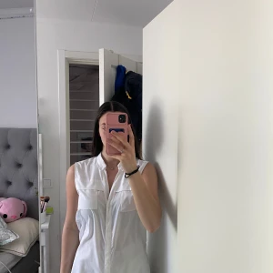 Vit ärmlös skjorta från Åhléns - Säljer en klassisk vit ärmlös skjorta från Åhléns i storlek 38. Skjortan har krage, knappar framtill och två bröstfickor. Perfekt till varma dagar och enkel att styla med olika outfits.