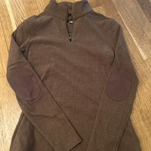 Brun tröja med dragkedja från HKM - Säljer en brun tröja från HKM med dragkedja och förstärkta armbågar. Perfekt för kyliga dagar med sin stilrena design och bekväma passform. Tröjan har långa ärmar och en hög krage.