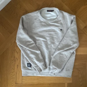 Grå sweatshirt från Morris - Säljer en stilren beige/grå sweatshirt från Morris med ett diskret emblem på bröstet. Tröjan har långa ärmar och en rund halsringning. Perfekt för en avslappnad stil.