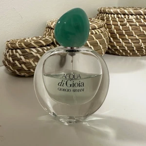 Acqua di Gioia från Giorgio Armani - Säljer en elegant parfymflaska av Acqua di Gioia från Giorgio Armani. Flaskan är rund och genomskinlig med en stilren design. Den har en unik grön kork som ger en fräsch känsla. Perfekt för den som älskar friska och naturliga dofter.