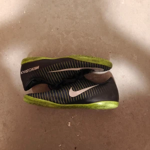 Svarta fotbollsskor från Nike - Säljer ett par svarta Nike fotbollsskor med vita detaljer och gröna sulor. Skorna har snörning och är perfekta för inomhusfotboll. De har en stilren design med Nike-loggan på sidan.