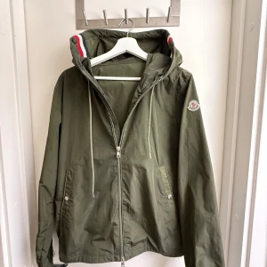 Grön vindjacka från Moncler - Snygg grön vindjacka från Moncler med huva och dragkedja. Jackan har långa ärmar och praktiska fickor med knappar. Perfekt för blåsiga dagar.Den har även Qr-kode på märket som man kommer in på Monclears hemsida (priset kan diskuteras)