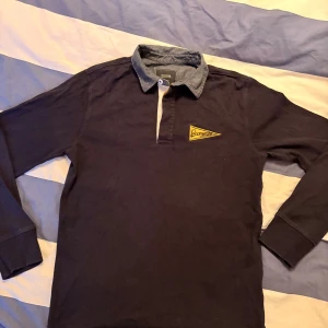 Mörkblå pikétröja från Boomerang - Säljer en stilren mörkblå pikétröja från Boomerang med en kontrasterande krage i ljusare blått. Tröjan har långa ärmar och en broderad logga på bröstet. Perfekt för en avslappnad men snygg look.