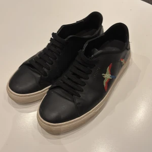 Svarta sneakers från Axel Arigato - Snygga svarta sneakers från Axel Arigato med broderade fåglar i färgglada nyanser på sidorna. Skorna har en klassisk design med snörning och en vit sula som ger en stilren look.