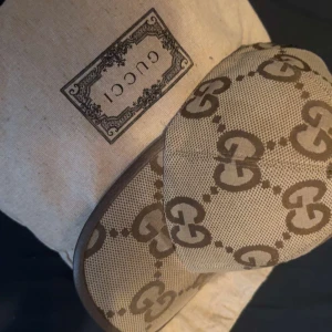 Beige keps från Gucci -  Äkta, Snygg beige keps från Gucci med det ikoniska GG-mönstret i brunt. Kepsen har en klassisk form och är tillverkad i ett slitstarkt material. Perfekt för att ge din outfit en lyxig touch, finns självklart kvitto och påse till den