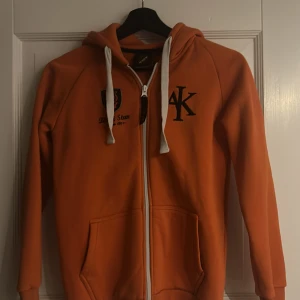 Orange hoodie med dragkedja från AIK Shop  - Säljer en orange hoodie med dragkedja från Aik. Tröjan har en broderad logga på bröstet och vita snören i huvan. Perfekt för en avslappnad stil.
