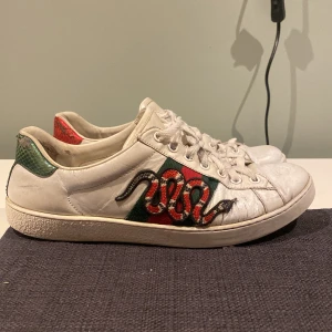 Gucci skor - Snygga vita sneakers med ett coolt ormmönster på sidan. Skorna har gröna och röda detaljer samt snörning. Perfekta för en trendig look.