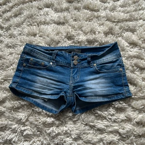 Lågmidjade jeansshorts  - Snygga blå jeansshorts från M-Cherry med dekorativa knappar framtill och klassiska femficksdesign. Perfekta för en avslappnad stil med en touch av edge. De har en lågmidjad passform och är idealiska för varma sommardagar. Priset går att diskutera.