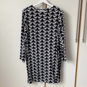 Svartvit klänning med kattmönster - Cool svartvit klänning med ett kattmönster. Klänningen har långa ärmar och rak modell. Sval och mjuk i 100% viskos. Djup knäppning på ryggen med två knappar. Längd 92 cm mått runt höfterna 104 cm. 