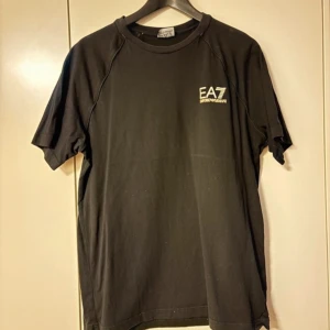 Svart t-shirt från EA7 - Säljer en stilren svart t-shirt från EA7 med korta ärmar och en liten logga på bröstet. Perfekt för en casual look eller träning. Skön passform och enkel design.