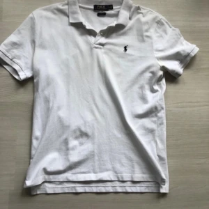 Vit pikétröja från Polo Ralph Lauren - Säljer en klassisk vit pikétröja från Polo Ralph Lauren med den ikoniska broderade loggan på bröstet. Tröjan har korta ärmar och en stilren krage med knappar. Perfekt för en avslappnad och stilren look.