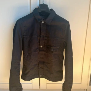 Svart overshirt från J.Lindeberg - Snygg svart jeansjacka/overshirt från J.Lindeberg med klassisk krage och knappar framtill. Jackan har en bröstficka med knapp och långa ärmar. Perfekt för en stilren look.