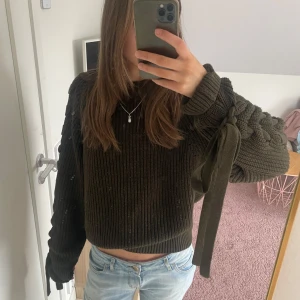 Olivgrön stickad tröja med snörning - Säljer en olivgrön stickad tröja med unika snörningsdetaljer längs ärmarna. Tröjan har en croppad passform och långa ärmar, perfekt för en trendig look. Den är både mysig och stilren, idealisk för kyligare dagar. Har två hål för tummarna 