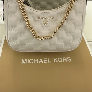 Vit axelväska från Michael Kors - Snygg vit axelväska från Michael Kors med quiltad design och guldfärgad kedja. Väskan har en dragkedja upptill och är perfekt för att bära dina nödvändigheter med stil. Den eleganta designen gör den till ett måste i garderoben.