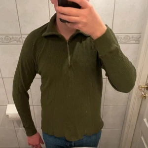 Olivgrön ribbad tröja med dragkedja - Säljer en olivgrön ribbad tröja med dragkedja. Tröjan har långa ärmar och en hög krage som ger en stilren look. Perfekt för kyligare dagar. Passar bra till jeans eller chinos.