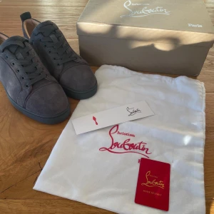 Grå sneakers från Christian Louboutin - Snygga grå sneakers från Christian Louboutin med den ikoniska röda sulan. Skorna har snörning och är tillverkade i skinn. Perfekta för dig som vill ha en stilren look med en touch av lyx.