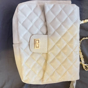 Grå väska  - Snygg grå axelväska med quiltat mönster och guldfärgad kedja som axelrem. Väskan har en praktisk dragkedja och ett elegant spänne framtill. Perfekt för att ge en stilren touch till din outfit.
