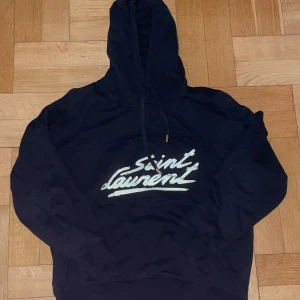 Svart Saint Laurent hoodie🔥 - Säljer en snygg svart hoodie från Saint Laurent med deras ikoniska logga i vitt/kräm färg på framsidan. Perfekt för en stilren och avslappnad look. Hoodien har en klassisk passform med långa ärmar och justerbar huva. Såklart jälvigt snygg.🔥🔥🔥Använd ett få par gånger och tvättad ungefär 2-3 gånger.