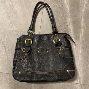 Svart axelväska med gulddetaljer - Snygg svart axelväska i syntetmaterial med guldfärgade detaljer och dragkedjor. Väskan har två handtag och flera praktiska fack både inuti och utanpå🥰