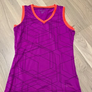 Lila tennis sett från TecnoPro - Säljer ett lila tennis sett från TecnoPro med geometriskt mönster och orange detaljer. Tennis setter är ärmlöst och har en v-ringad hals. Perfekt för träning med sitt DRY PLUS-material som håller dig torr.