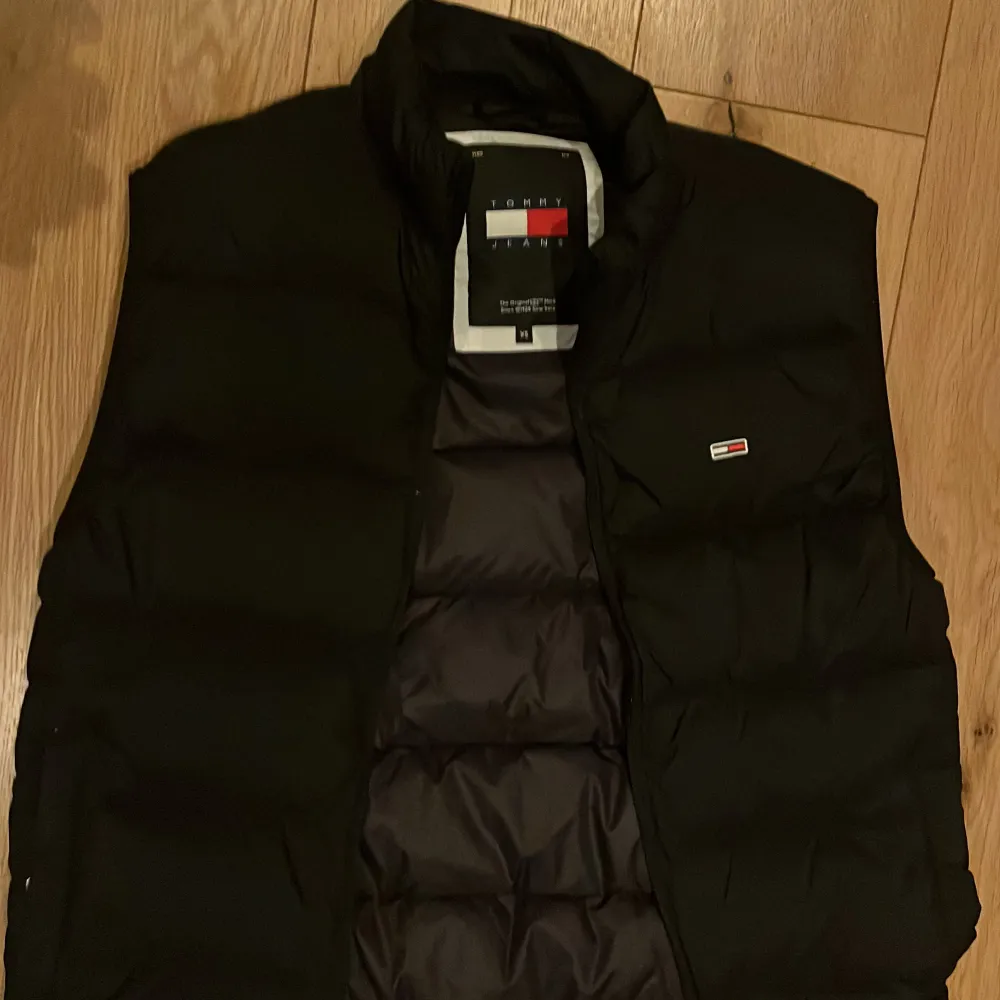 Snygg svart dunväst från Tommy Hilfiger med en stilren design. Västen har en diskret logotyp på bröstet och en hög krage för extra värme. Perfekt för lager-på-lager under kyliga dagar.. Takit.